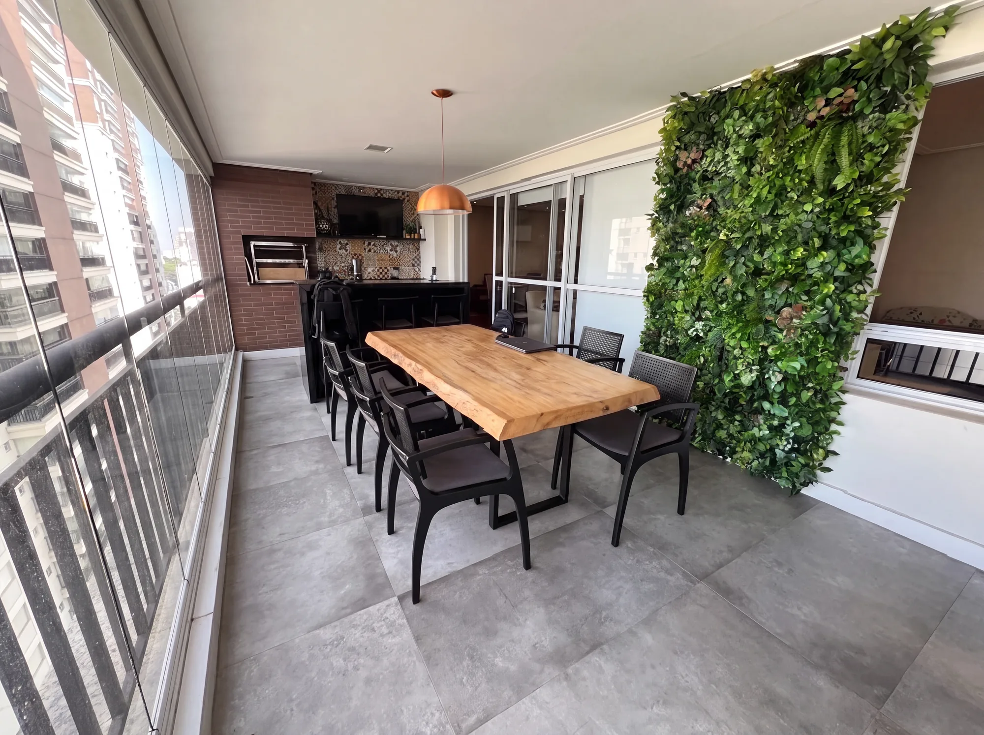 Varanda gourmet contínua da Casa das Caldeiras: mesa de madeira maciça sob pendente de cobre, churrasqueira de alvenaria em tijolo aparente ao fundo e jardim vertical à direita.