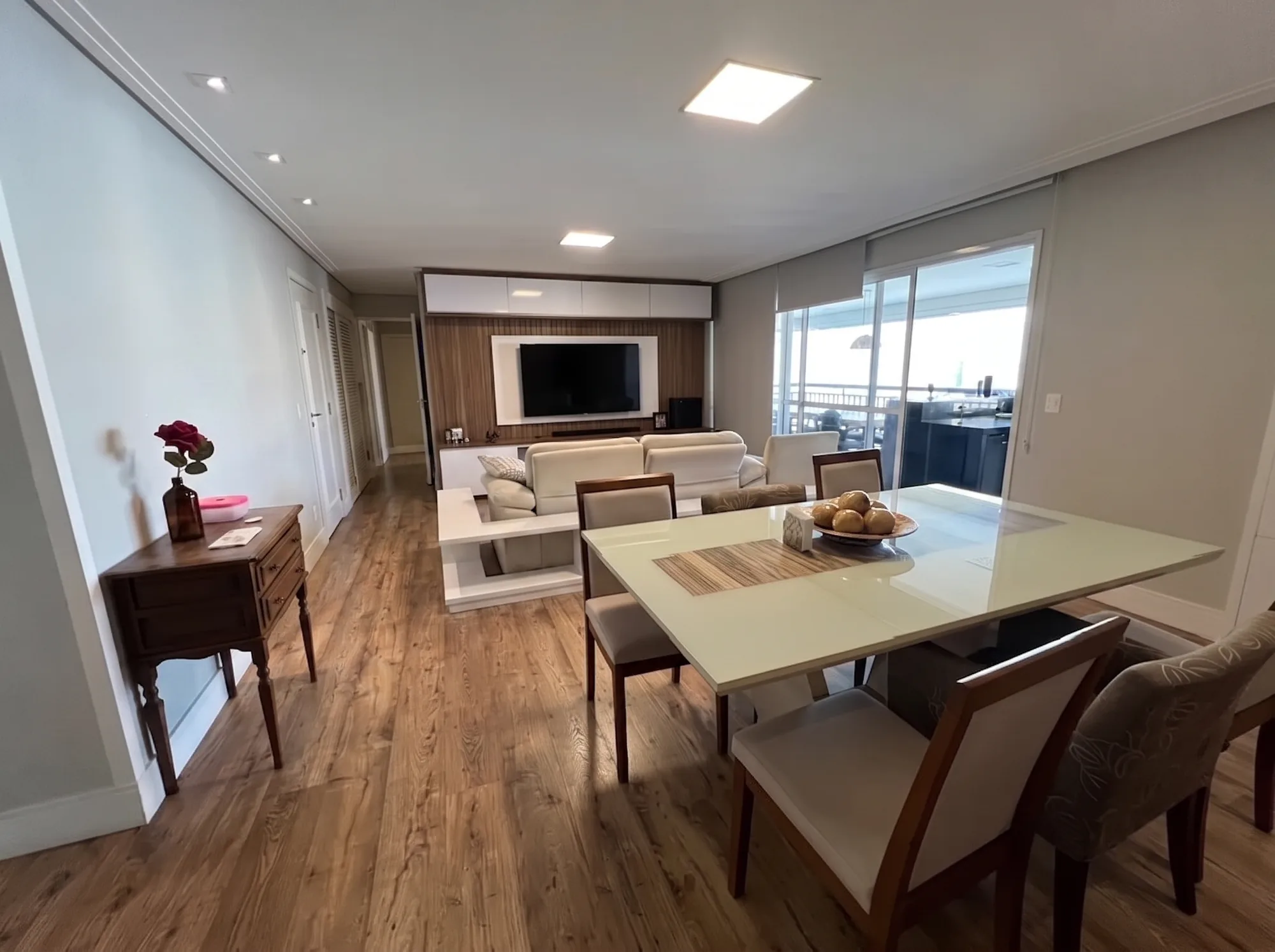 Sala em L com piso laminado tom mel, sanca e rodapé brancos e porta de correr de vidro em toda a extensão para a varanda.